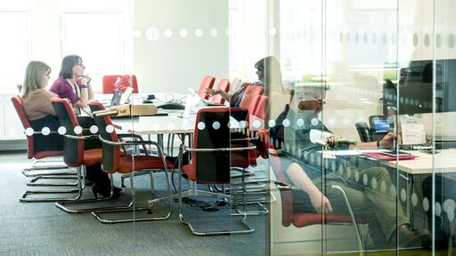 A picture of two office meeting rooms behind some floor to ceiling glass. One meeting room is straight on to the camara and has three individuals sitting at a long table and sitting on red chairs. The other meeting room is to the right of the camara and is quite obscured, but you can make out an individual seating on a red chair.