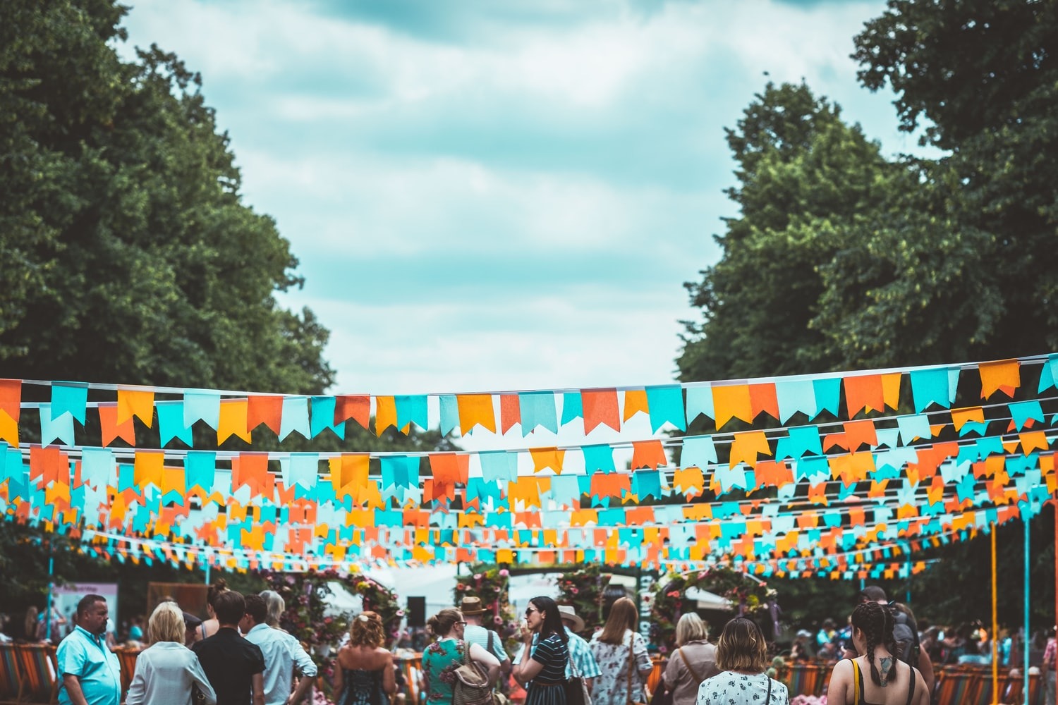 A picture of a crowd of people at a fate. Above the crowd of people is a continuous string of bunting suspended above their heads like a canopy of bunting. To the left and right of the crowd of people are very tall and luscious trees in lines.