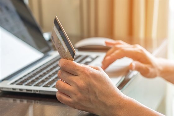 A picture of someone's lower arms with them sitting down at a table with a laptop in front of them. Their left lower arm is rested on the table and is holding a gold card or debit card. The face of this card is pointing towards them. Their right hand is on the laptop and is using the scroll pad. It appears that the person is reading the gold credit or debit card details and it about to enter them via their laptop.