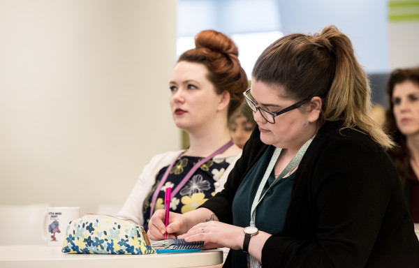 A photograph of two individuals sitting at a desk listening to a talk. The individuals are side on to the camara. You can only see the individuals from the waist up. One individual is writing and making notes.
