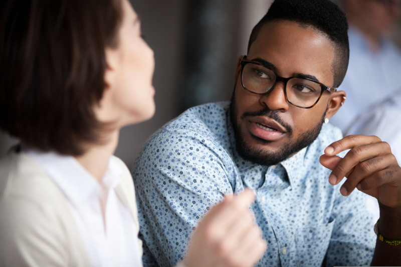 A photograph of two individuals holding a conversation. One individual is completely side-on from the camara with a blurred face and the other individual is slightly side-on but you can see their face clearly.